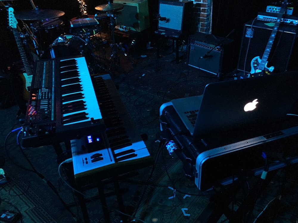 ライブ会場の床に置かれたシンセサイザーと、ケースの上で光るMacBook